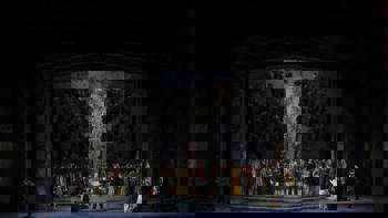 Photo from the main stage of the Greek National Opera during a performance in which there is a crowd of people gathered around a large dining table